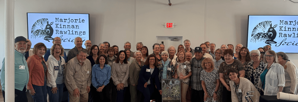A group of people stand indoors in front of Marjorie Kinnan Rawlings Society signs