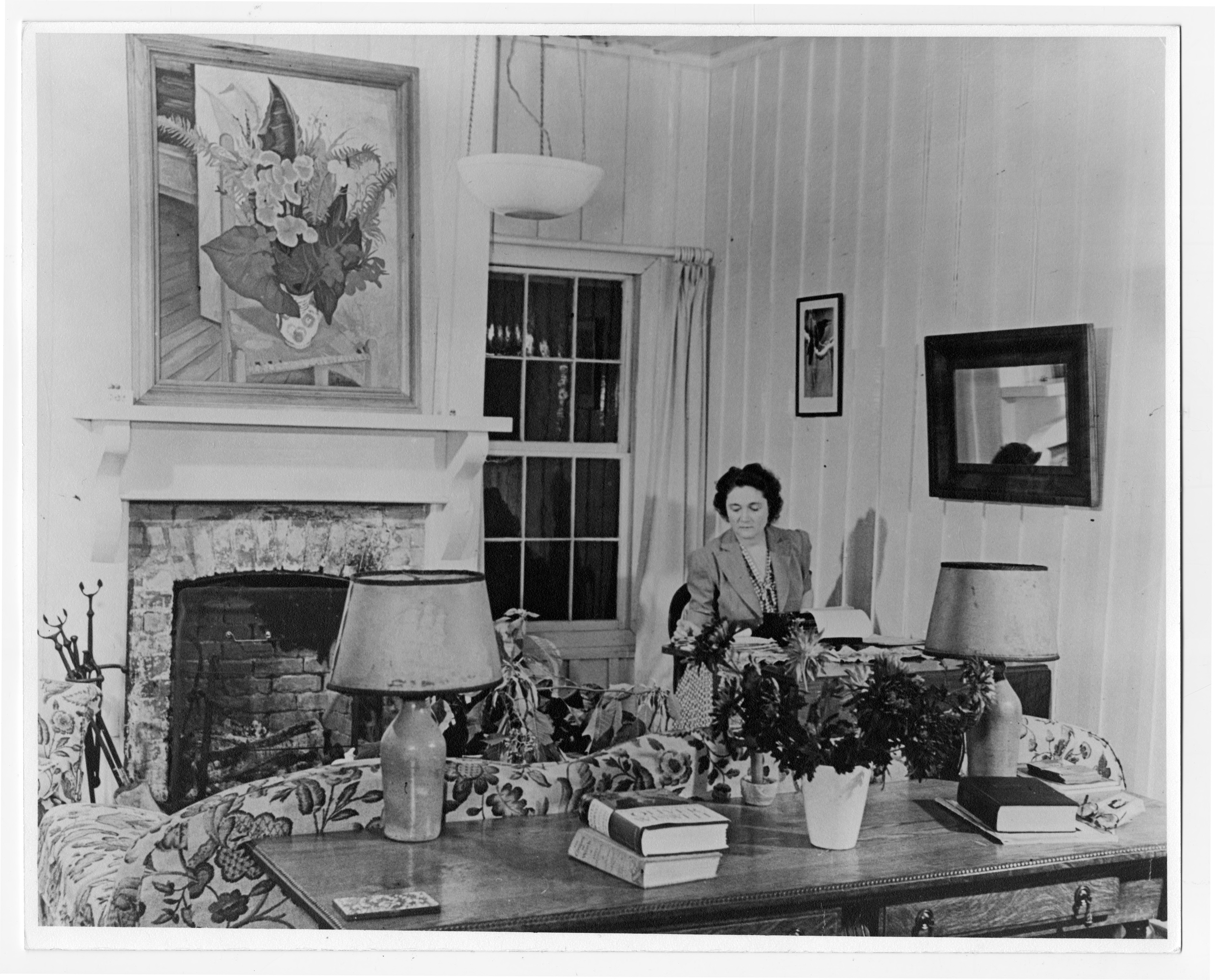 Marjorie Rawlings (middle aged adult) posed at writing table, south end of living room in her home. Painting over fireplace by Bob Camp. 